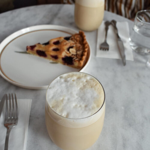 Two butterscotch lattes in glasses with foamed milk and a drizzle of butterscotch syrup on top. There is also a lslice of cherry lattice pie on a plate.