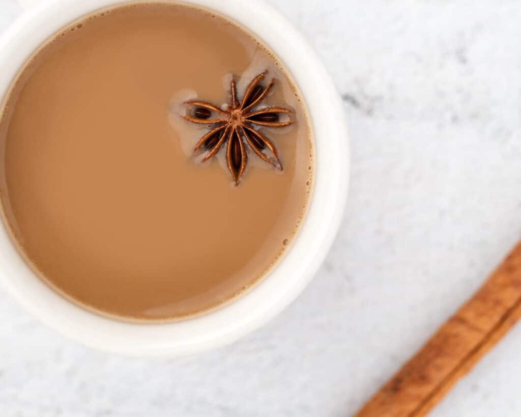 A chai latte in a white cup with a star anis spice floating on top. Its standing on a white surface with a cinnamon stick in the foreground