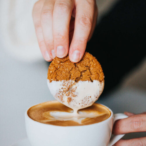 a white cup and saucer holding a gingerbread latte, with a hand dipping a ginger cookie into the latte.