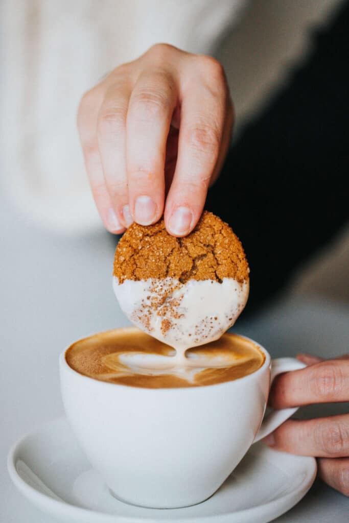 a white cup and saucer holding a gingerbread latte, with a hand dipping a ginger cookie into the latte.
