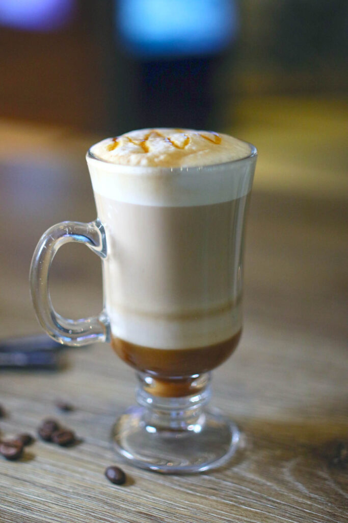Honey latte in a latte glass with creamy foam and a drizzle of honey on top, standing on a wooden table with some scattered coffee beans.