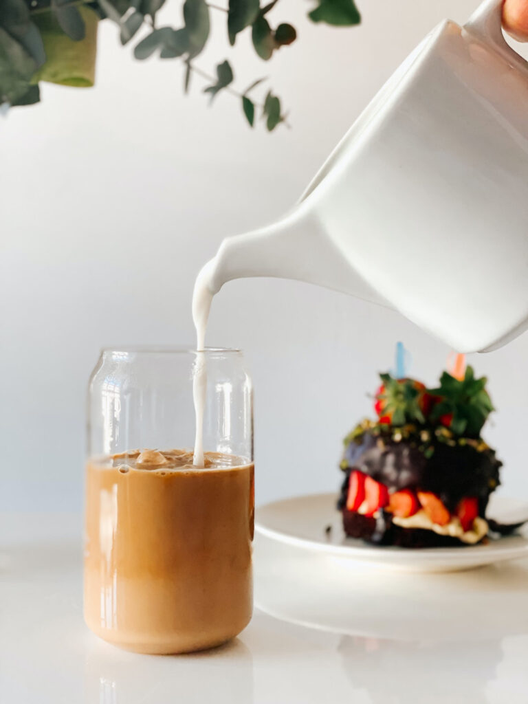 milk pouring from a white teapot into an iced blonde latte on a white surface and a fancy chocolate and strawberry cake in the background.