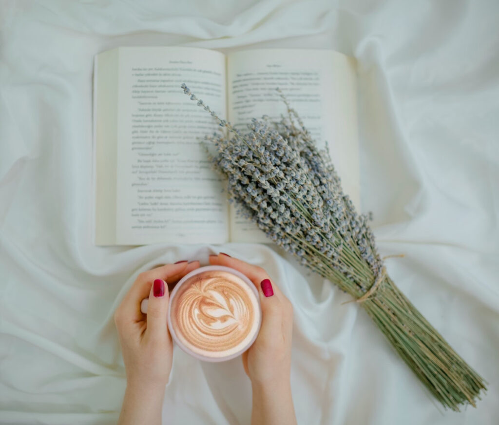 Two hands holding a mug of lavender latte resting on a white table cloth with a bunch of fresh-cut lavender nearby