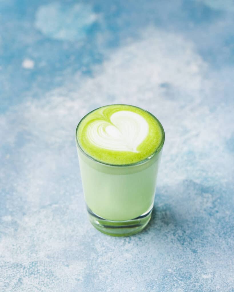 a green matcha chai latte in a glass with a latte art heart on top. Its standing on a blue and white surface styled like the sky.