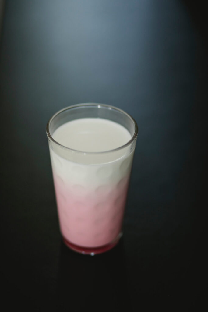 A glass of strawberry latte with a white foam top standing on a black surface.