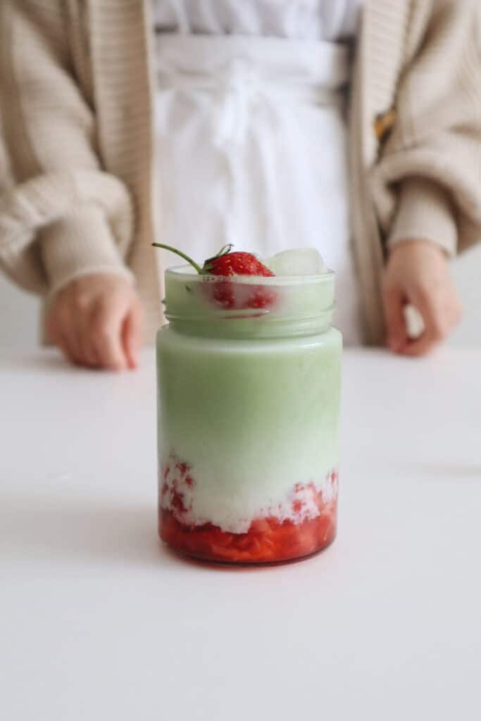A strawberry matcha latte with pulped strawberries at the bottom of the glass with matcha and milk above topped with a whole strawberry, standing on a white surface.