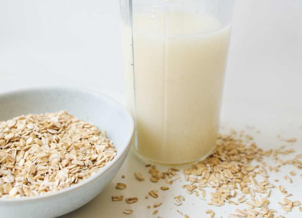 A tall glass of oat milk with a bowl of oats and oats scattered around. One of the best non-dairy milks for coffee.