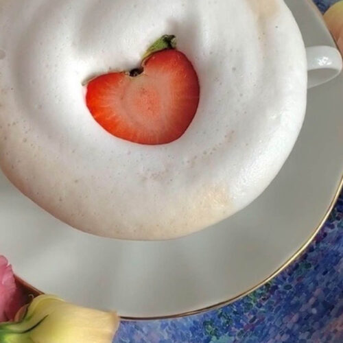Slice of strawberry on top of a frothy latte in a cup and saucer.