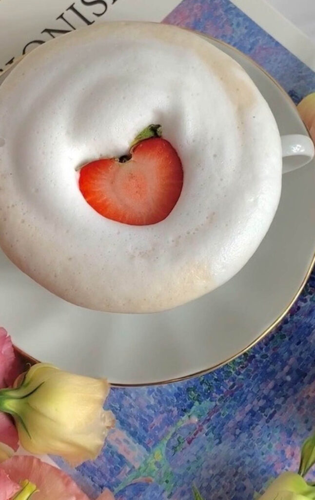 Slice of strawberry on top of a frothy latte in a cup and saucer.