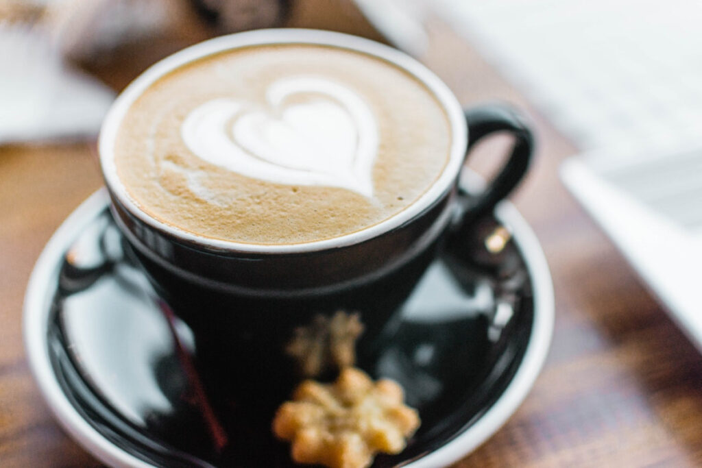 A black cup of vanilla oat milk latte, with a latte art heart design. The saucer holds a small cookie.
