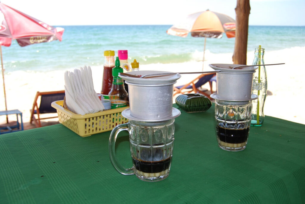 Two Vietnamese coffee filters brewing coffee, sitting on a green surface with a beach in the background.