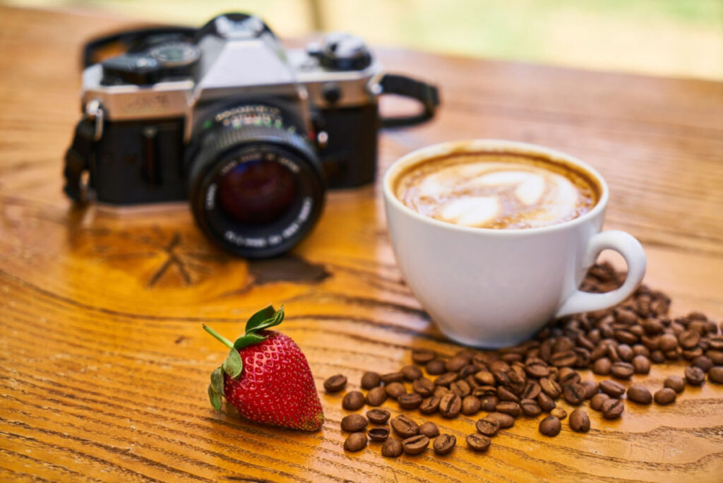 a cup of latte on a table with a strawberry, some coffee beans and a camera also sitting on the table.