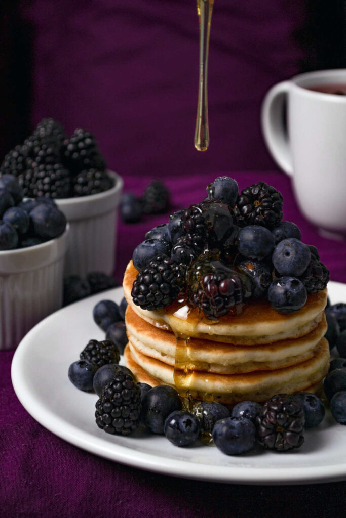 Agave syrup being poured over a stack of pancakes, which are covered in blueberries and blackberries.