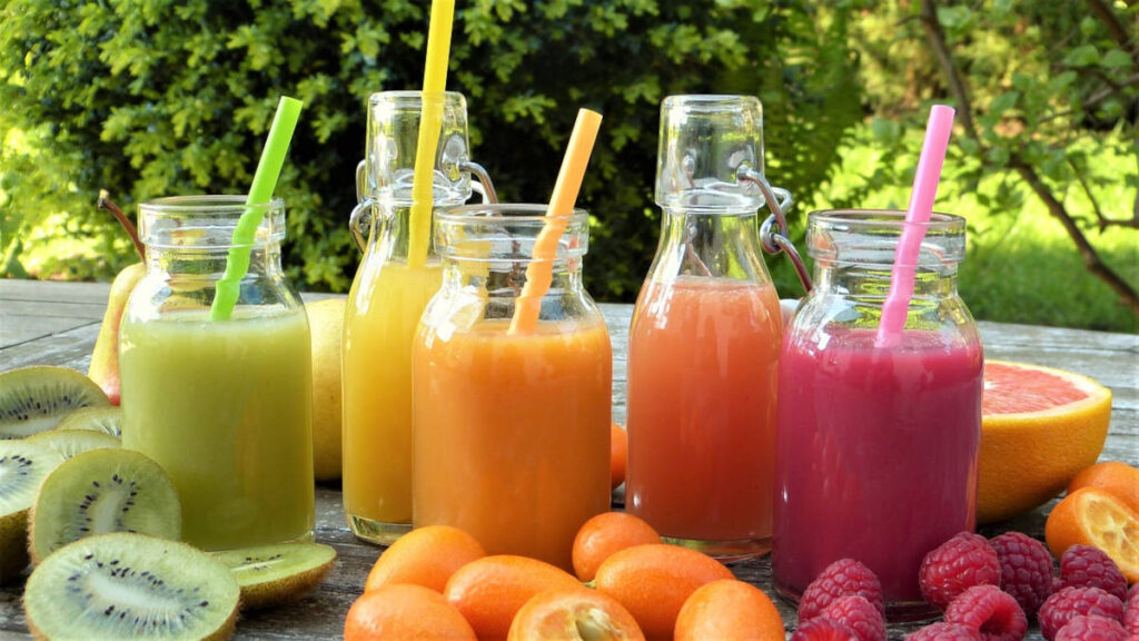 Five jars of different colored fruit juices, with straws. Surrounded by lots of different types of fruit.