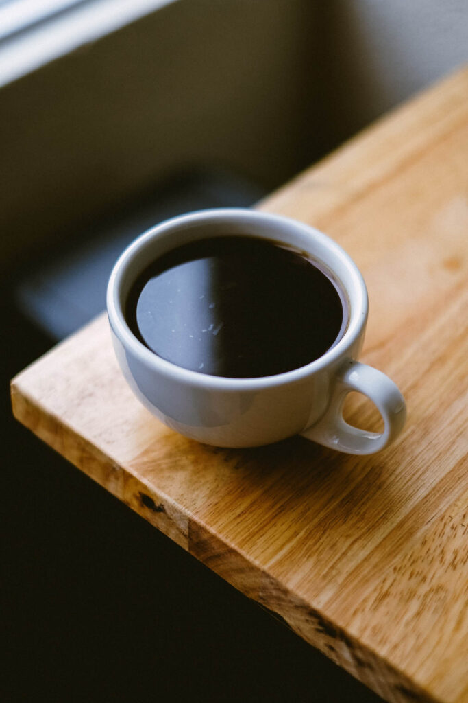 A cup of black coffee standing on the edge of a wood table.