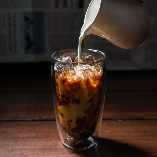 Chilled milk being poured over an iced black coffee in a glass.