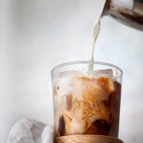 Chilled foamed milk being poured over 2 shots of espresso and ice in a glass.