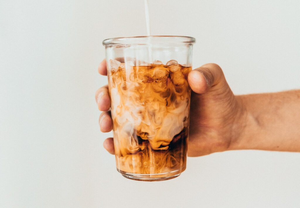 a hand wrapped around a glass of iced coffee, with a stream of milk pouring into the glass.