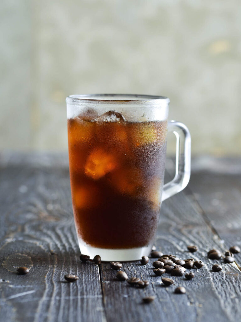 A frosty glass mug of iced black coffee standing on a piece of wood.